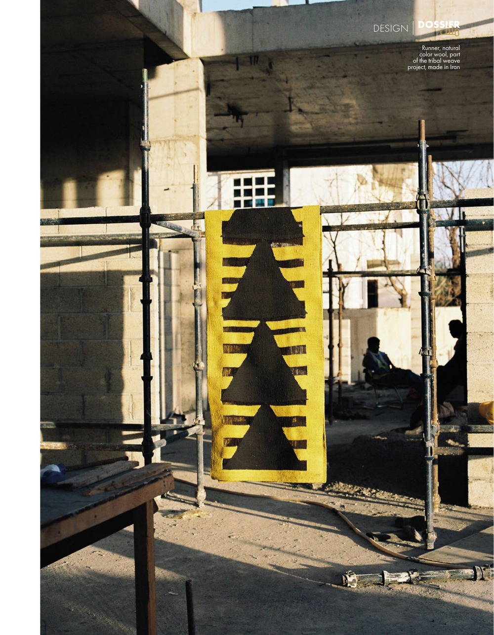 A yellow and black arrow-patterned runner from the Tribal Weave Project hanging from scaffolding, workers resting behind it