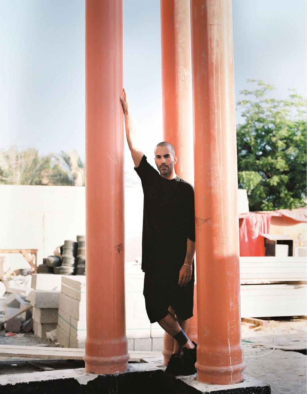 Taher Asad-Bakhtiari dressed in black, leaning against tall orange columns on a construction site